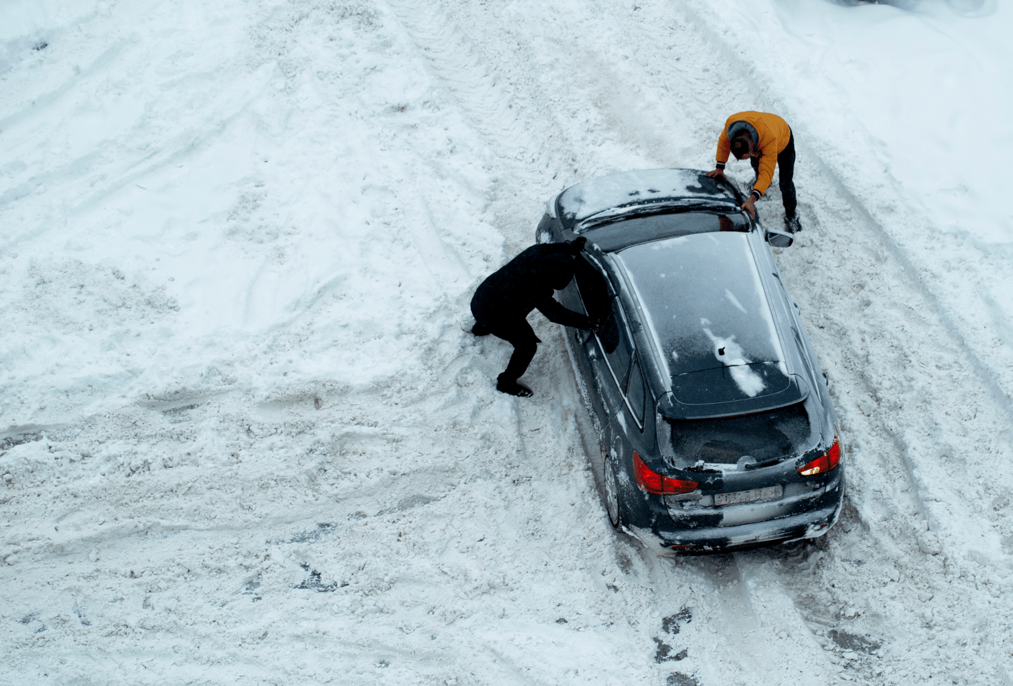 Car stuck in snowy roadway. Two people pushing car.